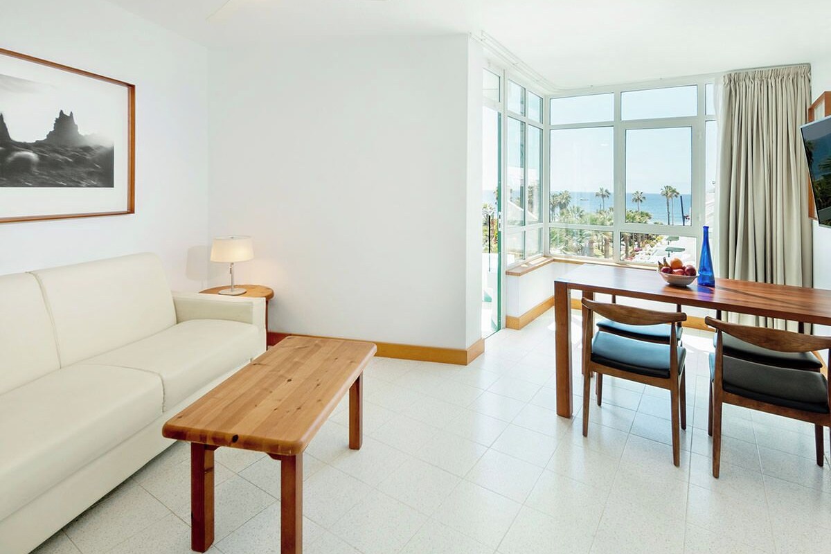 A hotel room with a white sofa, a wooden table, and a dining area with chairs. There is a large window with a view of the ocean and palm trees.