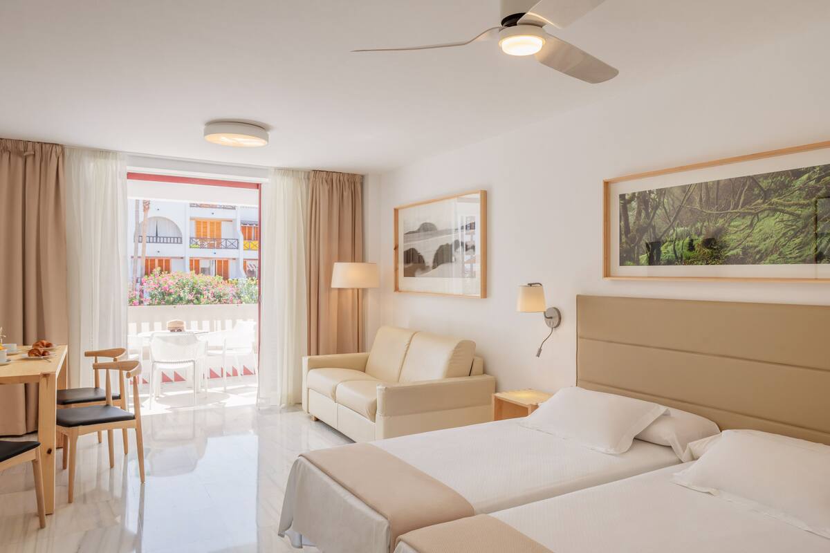 A hotel room with two beds, a sofa, a dining area, and a balcony with a view of buildings and flowers.