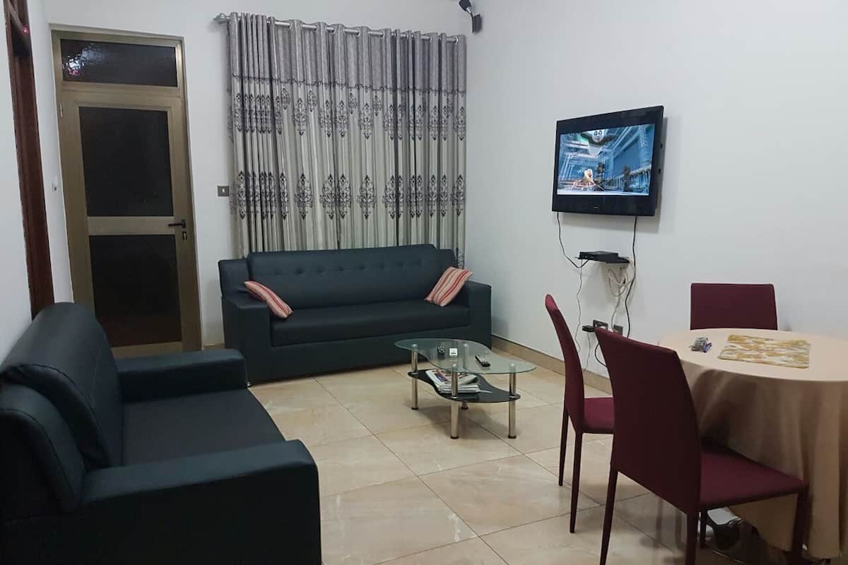 A living room with a sofa, a glass coffee table, a dining table with chairs, a television mounted on the wall, and a door with a curtain.