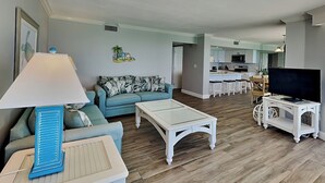 Sala de estar con sofá, una mesa de centro y un mueble para televisor. Se observa una cocina al fondo con una zona de comedor.