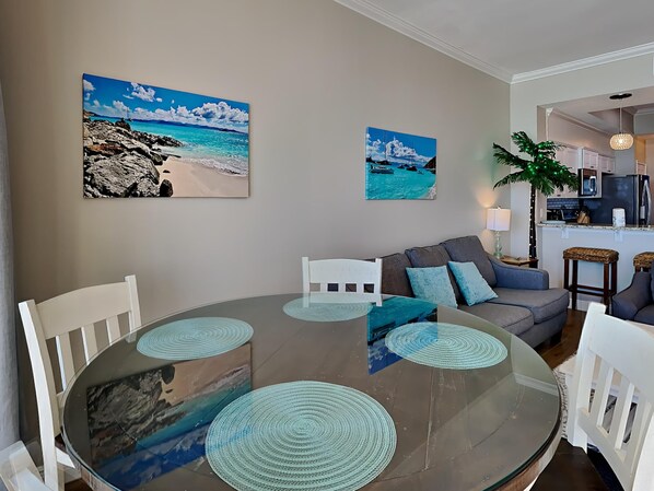 Ein Essbereich mit einem Glastisch und Stühlen, einem grauen Sofa und zwei an der Wand befestigten Bildern einer Strandansicht.