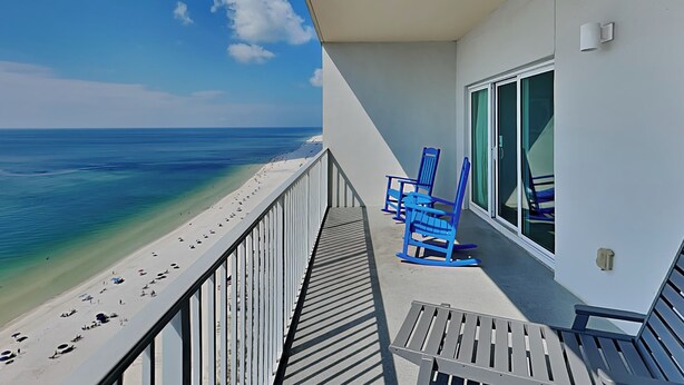 Een balkon met uitzicht op een strand en de oceaan. Er staan blauwe stoelen en een tafel op het balkon.