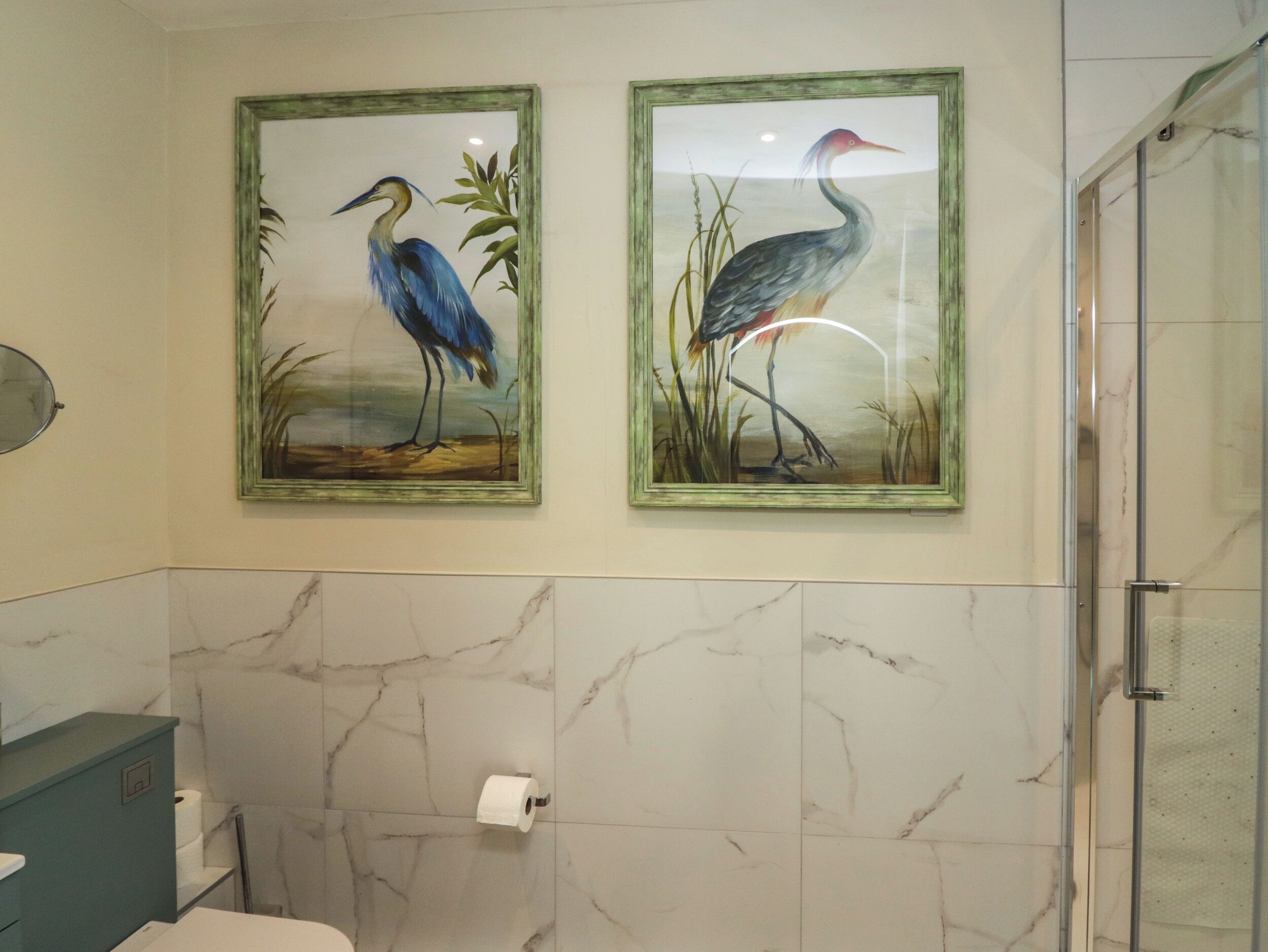 A bathroom with two framed paintings of birds, marble walls, a toilet, and a shower area.