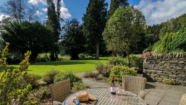 A round wicker table with a bottle and glasses, surrounded by wicker chairs, set on a patio with a stone wall and lush greenery in the background. - Lakeside View (Ulverston)