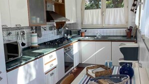 A compact kitchen with white cabinets, a stove, and a window with curtains. - Villetta a Castiglioncello (Rosignano Solvay-Castiglioncello)