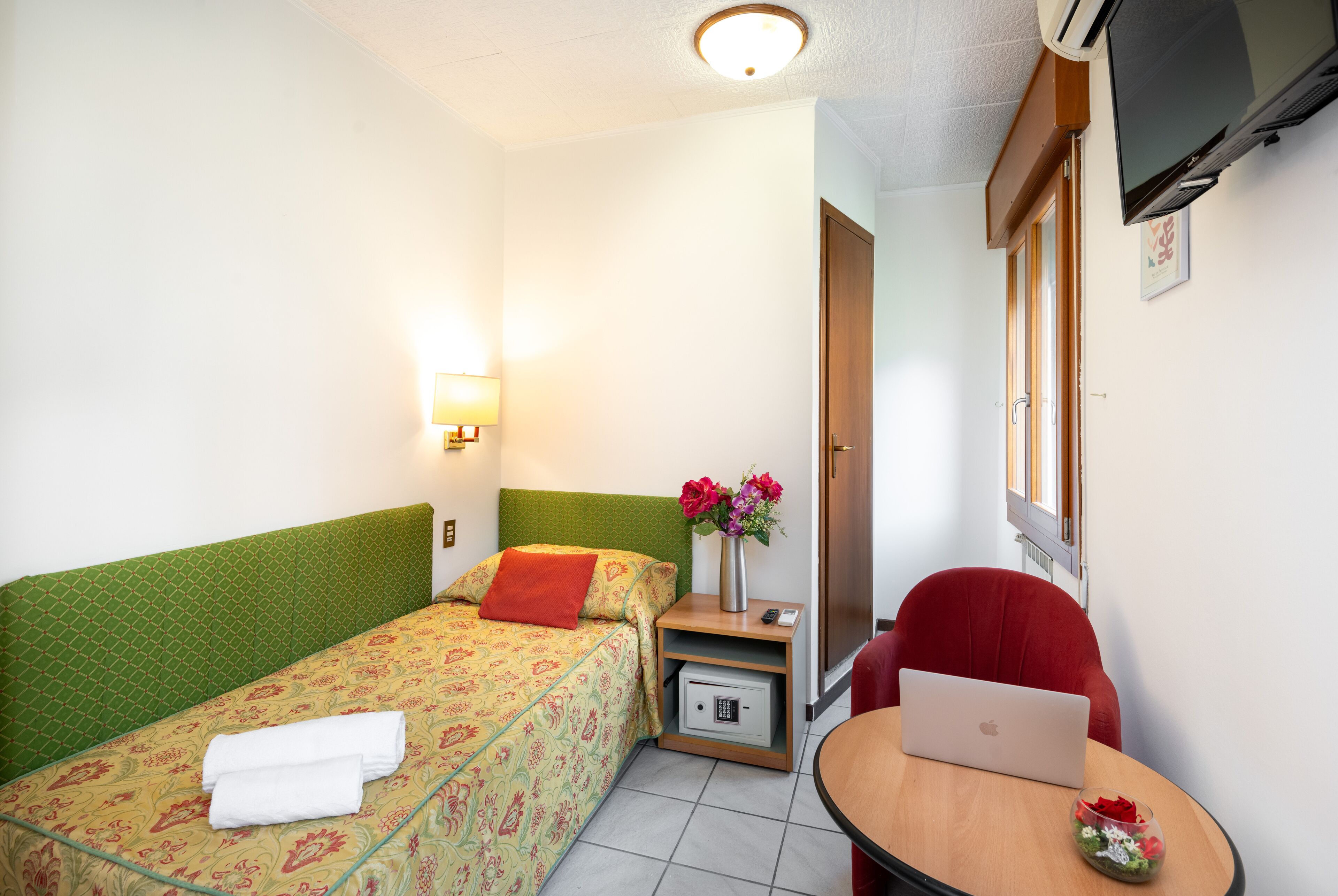 Ein Hotelzimmer mit Bett, Schreibtisch mit Laptop, Stuhl, Nachttisch mit Lampe und an der Wand montiertem Fernseher.