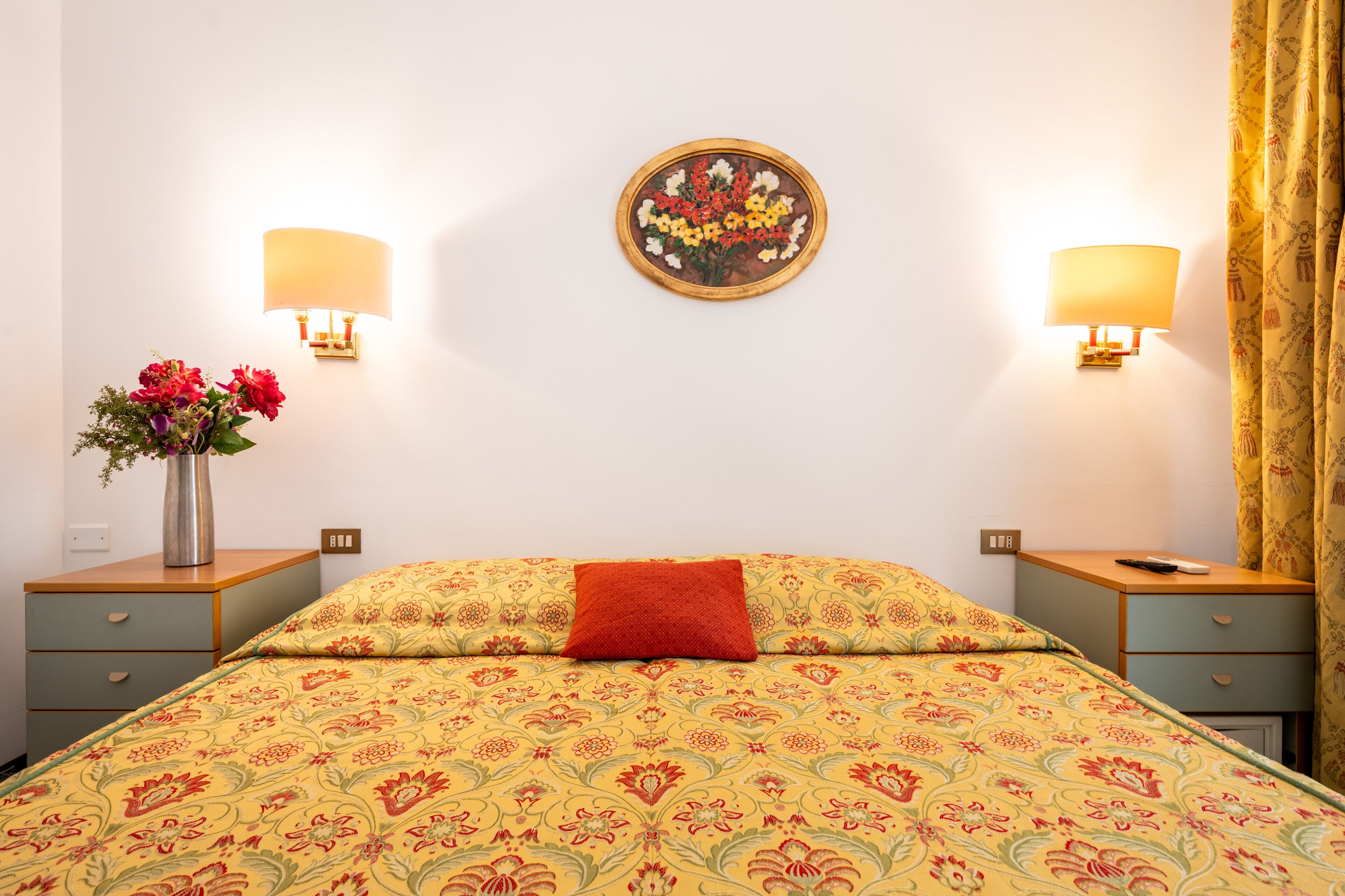 Una camera d'hotel con un letto, due comodini, un vaso con fiori e una lampada a parete.