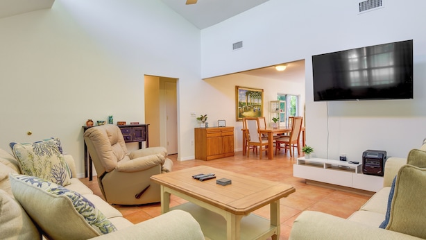 Una sala de estar con un sofá beige, una mesa de centro de madera, un televisor montado en la pared y una zona de comedor con una mesa y sillas de madera.
