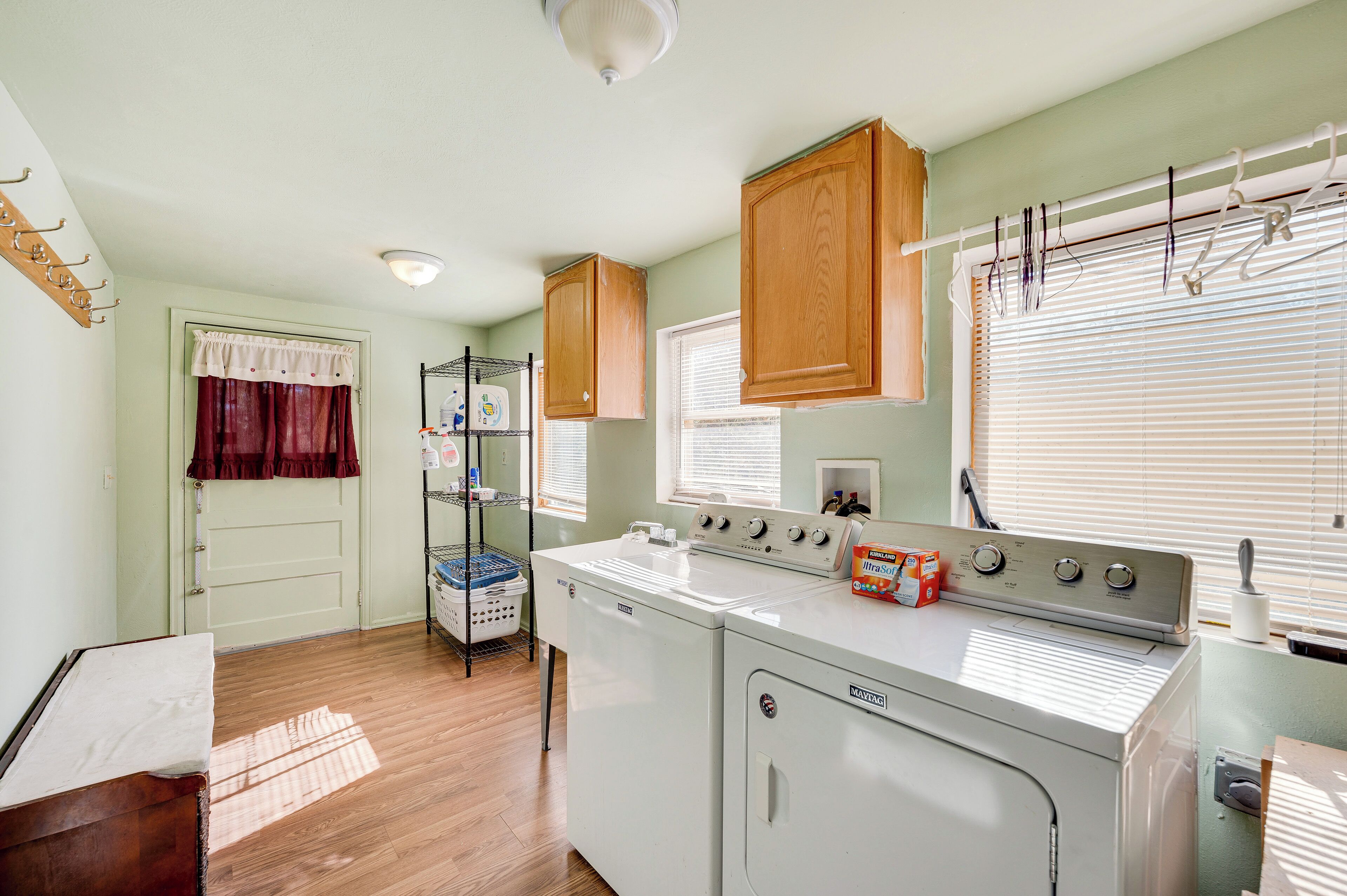 A laundry room with a front-loading washer and dryer, wooden cabinets, a drying rack, and a window with blinds.