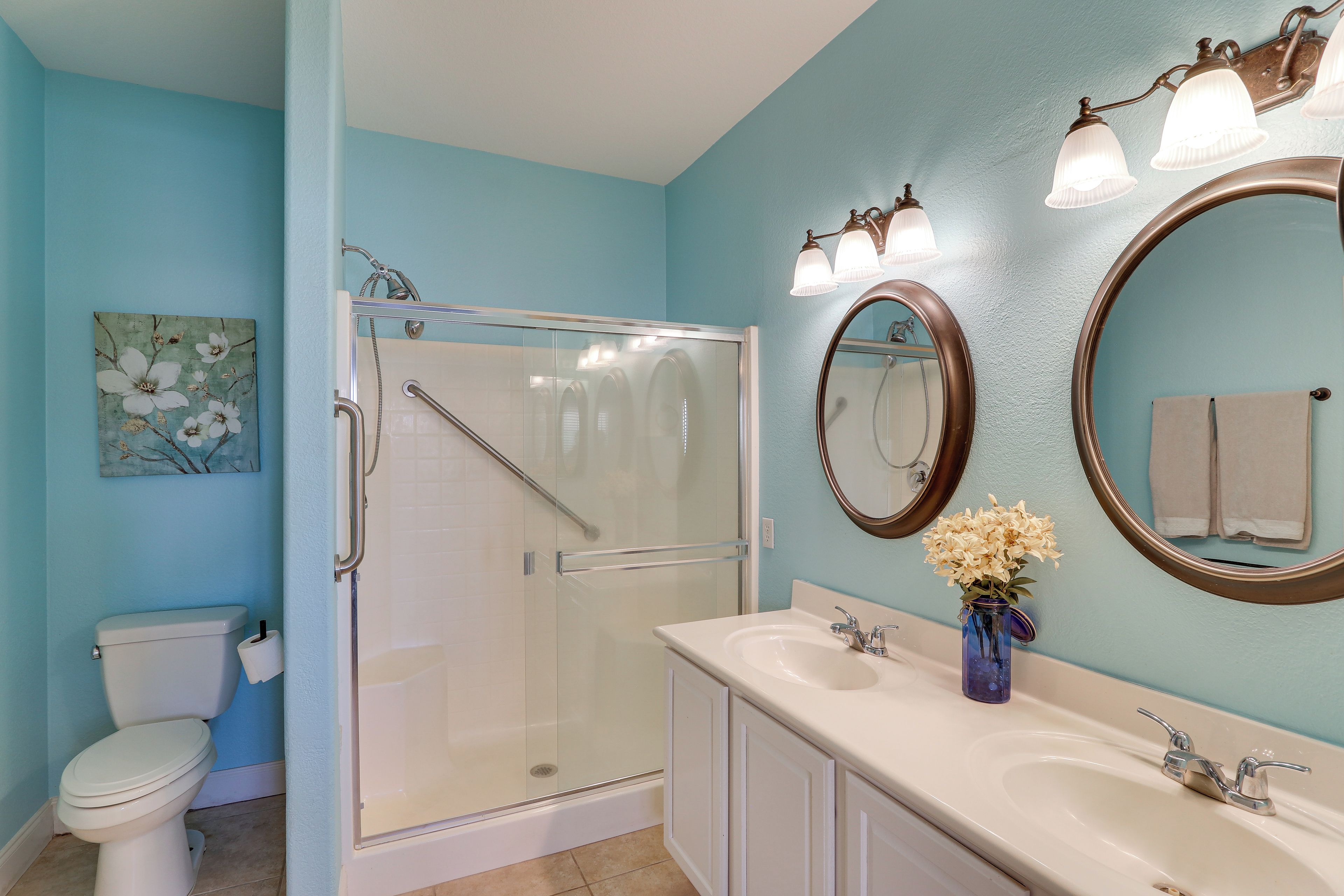 A bathroom with a toilet, a glass-enclosed shower, a sink with a vase of flowers, and two mirrors.