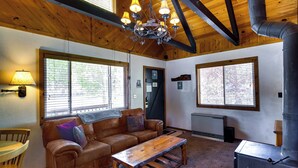 A cozy living room with a leather sofa, a wooden coffee table, and a chandelier. There are large windows providing natural light and a view of the outside. - Hot Tub & Deck: Big Bear City Cabin Near Skiing (Big Bear City)