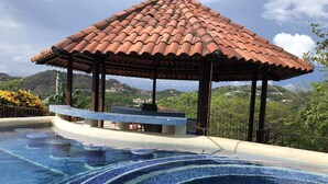 A swimming pool with blue mosaic tiles, a gazebo with a tiled roof, and a view of a hillside with buildings. - Breathtaking panoramic ocean view Penthouse Condo. (Playa Hermosa)