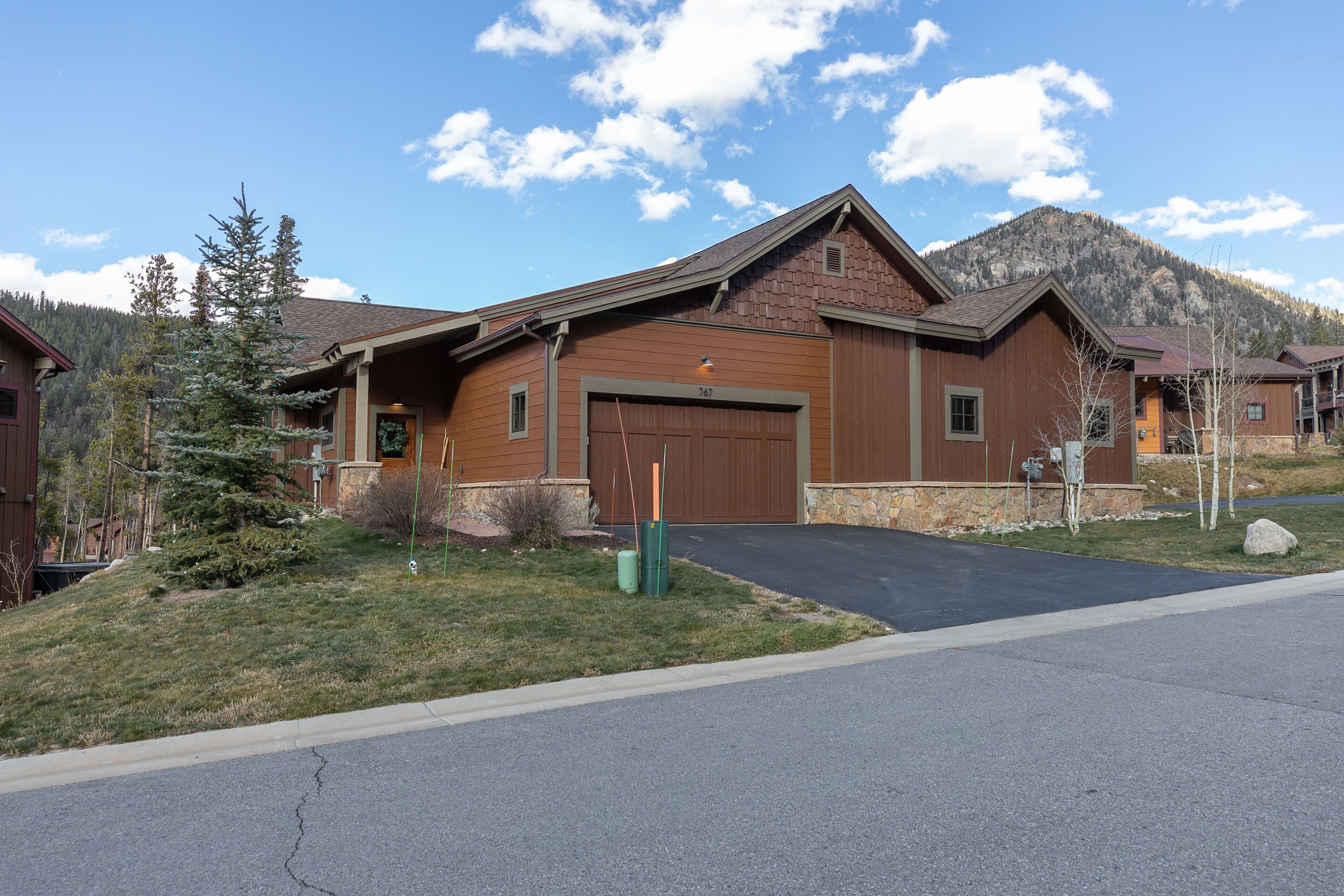 A two-story wooden house with a garage, surrounded by a grassy area and a mountain in the background.