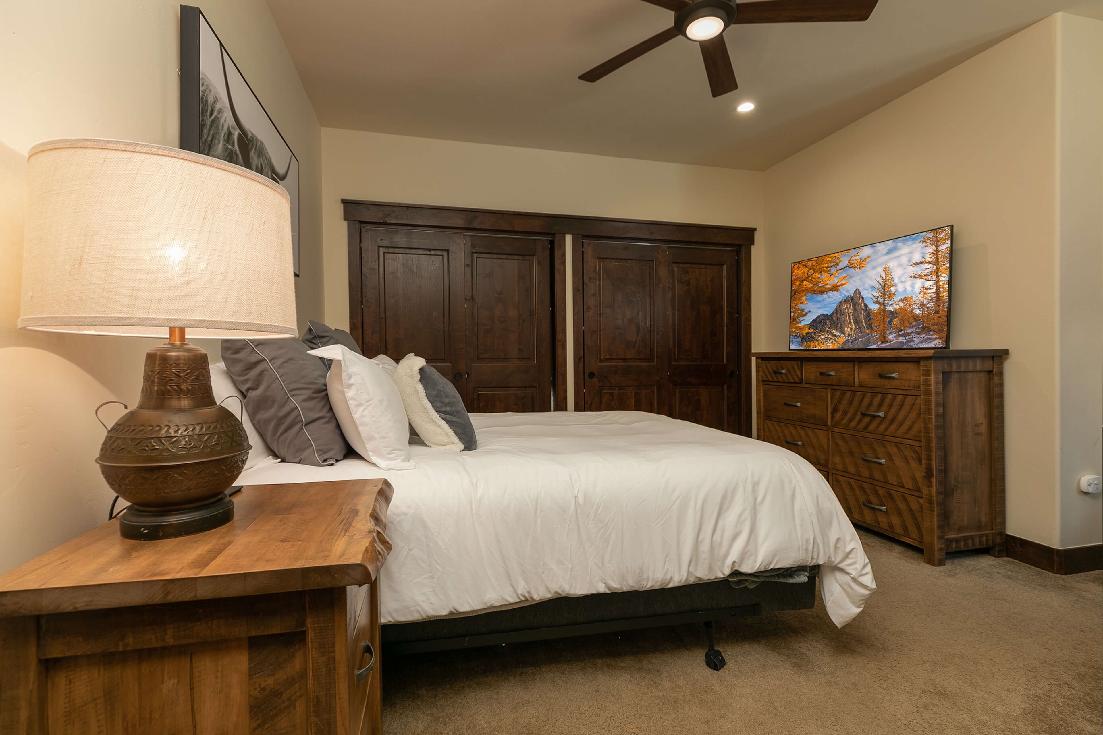 A bedroom with a bed, a wooden nightstand, a lamp, a ceiling fan, and a painting on the wall.