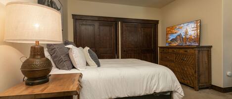 A bedroom with a bed, a wooden nightstand, a lamp, a ceiling fan, and a painting on the wall.