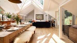 Un loft spacieux avec un plafond haut, un coin repas avec une table en bois dressée pour un repas, un canapé sombre et une grande fenêtre offrant une vue sur la verdure.