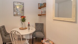 Een eetgedeelte met een ronde tafel, twee stoelen, een vaas met bloemen, een wijnfles en een wijnglas. Er is een boekenkast met boeken en een lijstspiegel aan de muur.