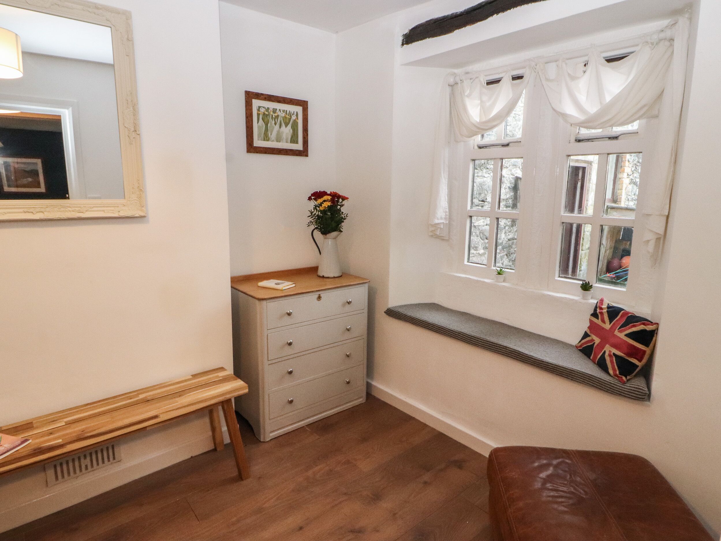 A room with a wooden bench, a white dresser, a window with white curtains, and a wall-mounted mirror.