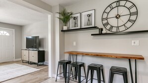 A modern kitchen with a wooden bar counter, four black stools, a large wall clock, and a potted plant.