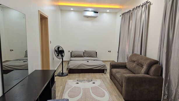 A bedroom with a bed, a sofa, a desk, a standing fan, and a rug. - Kams homes (Abuja)