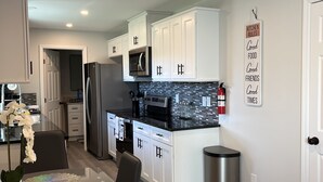 A modern kitchen with white cabinets, a stainless steel refrigerator, a microwave, and a black countertop. There is a kitchen sign on the wall and a trash can near the sink.
