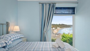 A bedroom with a bed, a nightstand, a lamp, and a view of the sea.