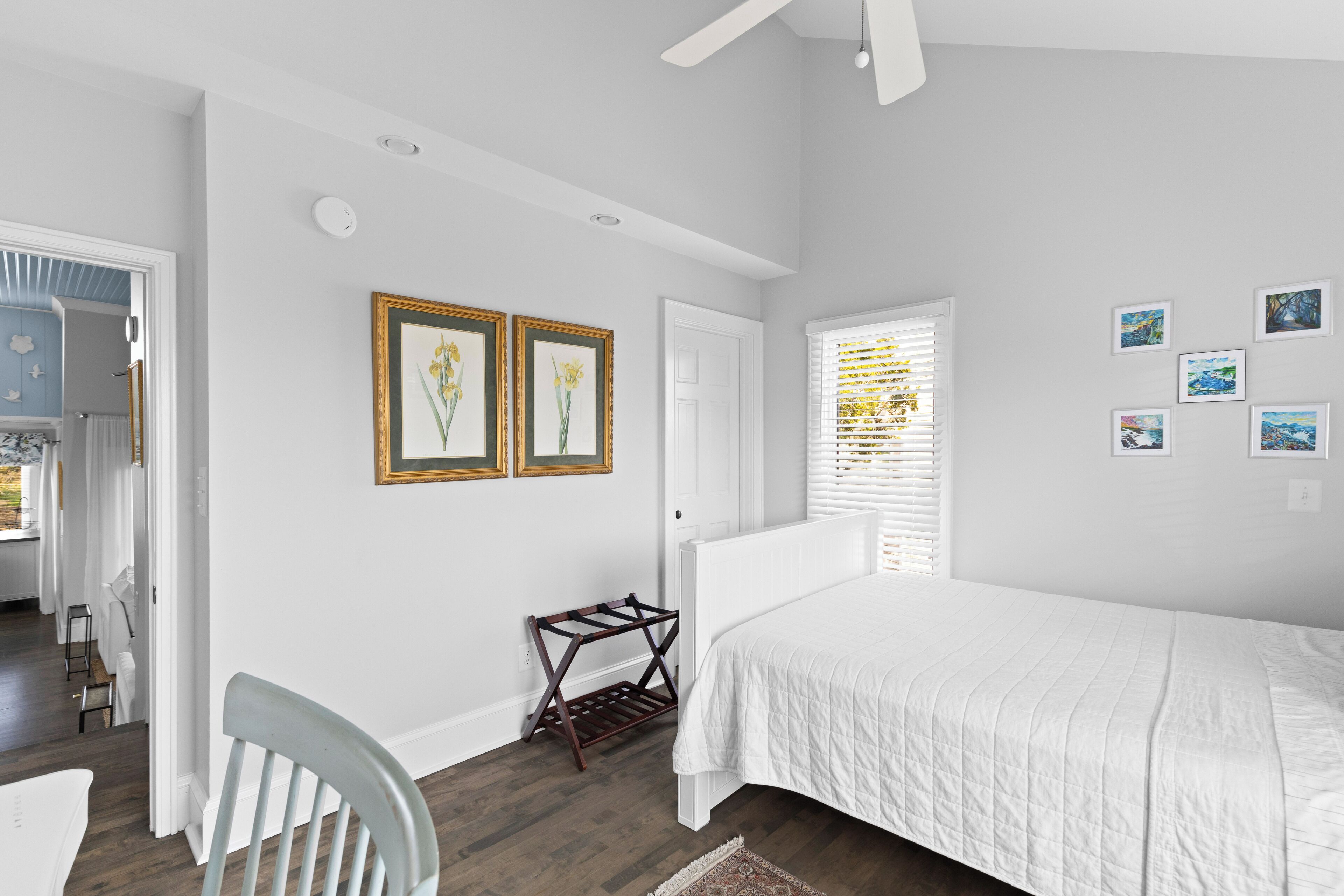 A bedroom with a large bed, a chair, a small table, and a window with blinds. There are framed pictures on the wall and a ceiling fan.