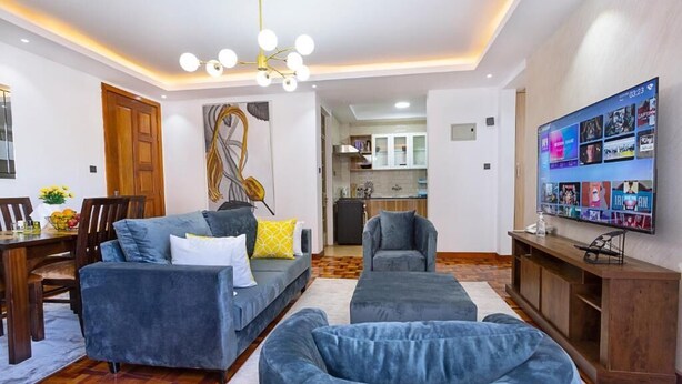 Ein modernes Wohnzimmer mit einer Couch, einem Hocker und einem an der Wand montierten Fernseher, der verschiedene Sender anzeigt.
