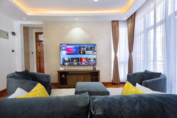 A modern living room with a large flat-screen TV mounted on the wall, two grey sofas with yellow cushions, and a wooden entertainment unit.