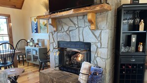 Una sala de estar acogedora con chimenea de piedra, un televisor fijado a una viga de madera y una estantería para vinos.