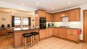 A modern kitchen with wooden cabinets, a central island with stools, a built-in oven, and a microwave. - Cairnhapple House (Stranraer)