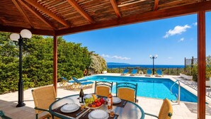 A covered outdoor area with a pool, dining table, and chairs, overlooking a scenic view. - Villa Thalia Katerina (Mousata)