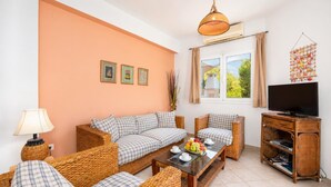A living room with a sofa set, a coffee table with fruit, a television on a wooden stand, and a wall-mounted air conditioning unit. - Villa Thalia Katerina (Mousata)
