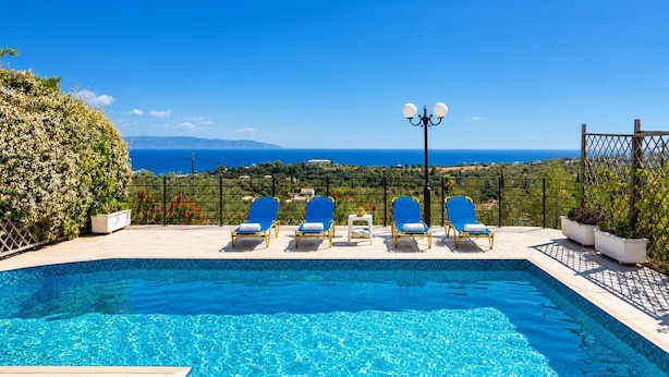 A pool with clear blue water, surrounded by lounge chairs and a lamp post, overlooking a scenic view of the sea and landscape. - Villa Thalia Katerina (Mousata)
