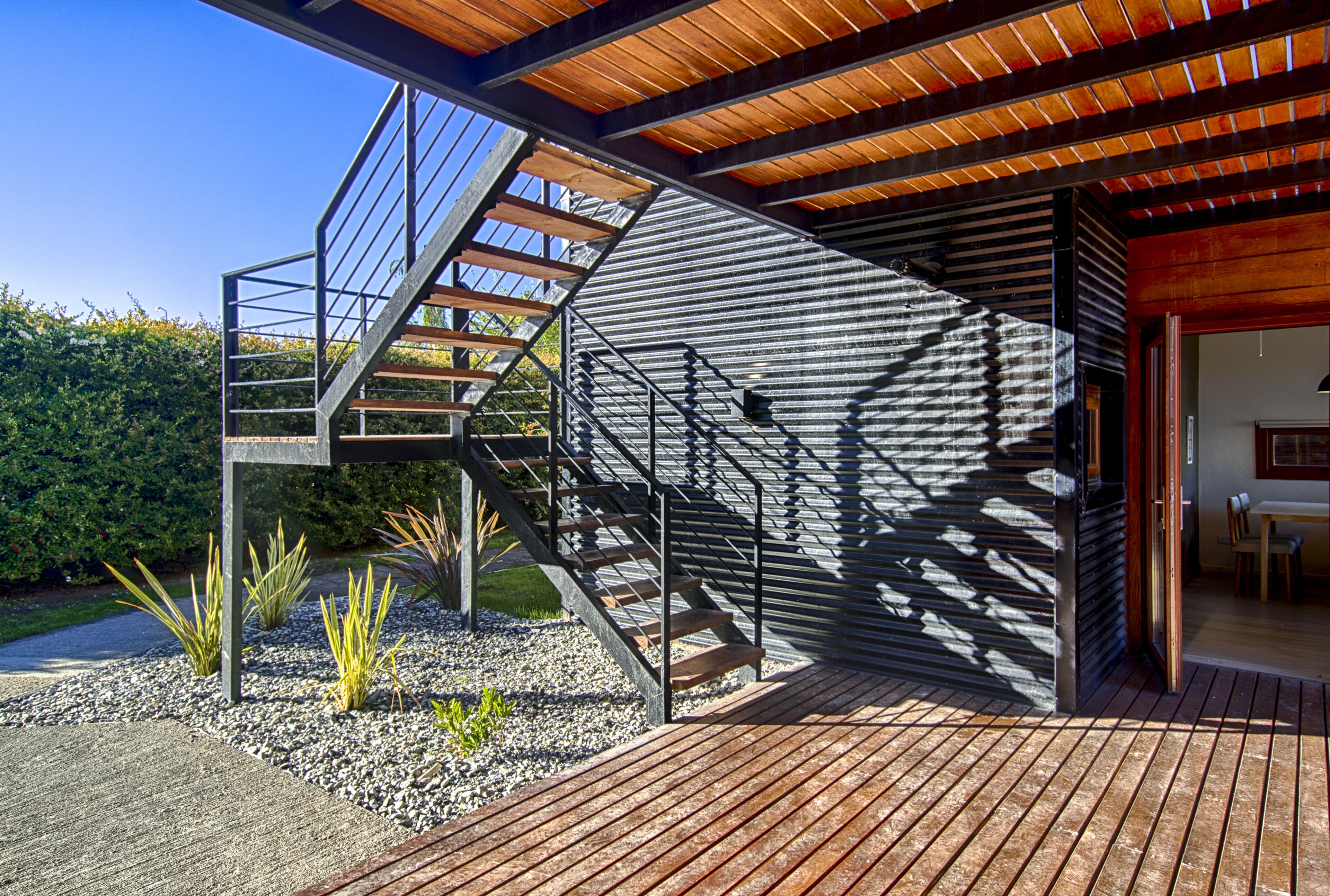 A modern outdoor staircase leading to a wooden deck with a view of a landscaped garden and a building with a sloped roof.
