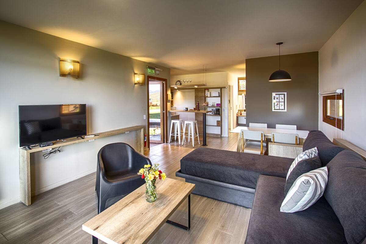 Una sala moderna con un televisor de pantalla plana, una mesa de centro de madera con un jarrón de flores, un sofá gris y un comedor con una cocina al fondo.