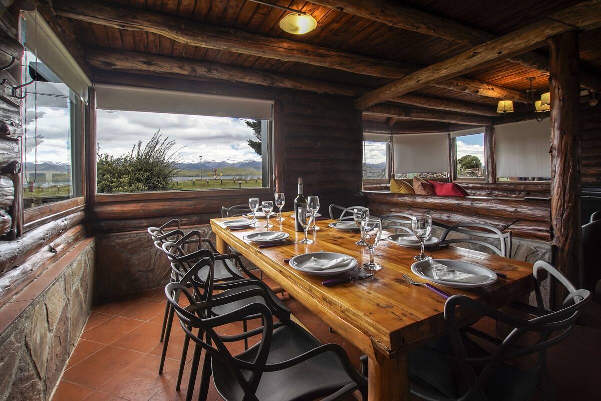 Una mesa de madera para comer dispuesta con platos, vasos y cubiertos, rodeada de sillas, con vistas a montañas a través de amplios ventanales.