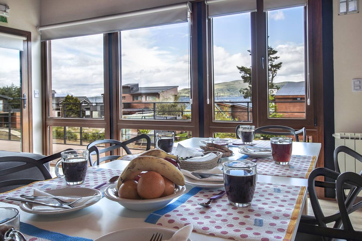 Una mesa de comedor con vistas al exterior, un bol de fruta y diversos platos y bebidas.