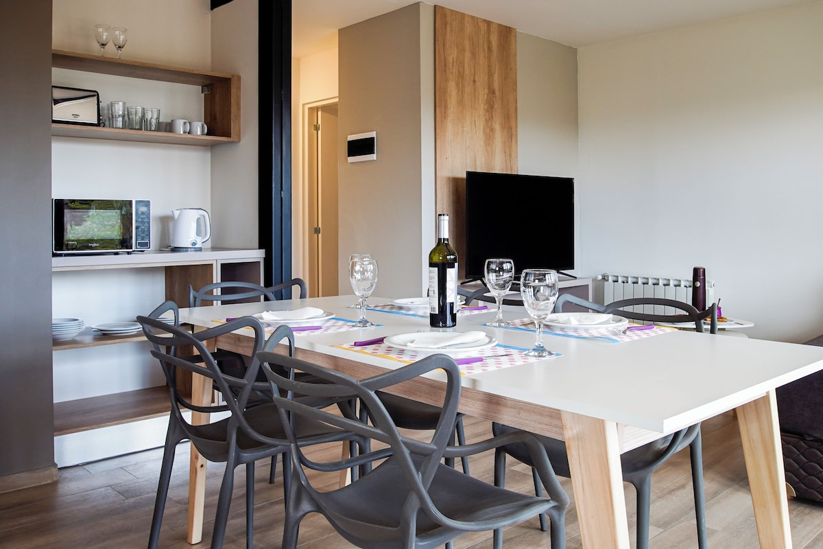 Un espace repas moderne avec une table blanche, des chaises grises et un téléviseur fixé au mur.