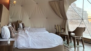 A spacious tented room with a large bed, a wooden nightstand, a chair, and a view of the outdoors.