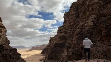 Une personne se tient sur un sentier rocheux dans un canyon, avec une vue sur des montagnes lointaines et un ciel nuageux.