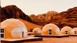 A row of dome-shaped tents with white curtains, set against a desert landscape with rocky hills in the background.