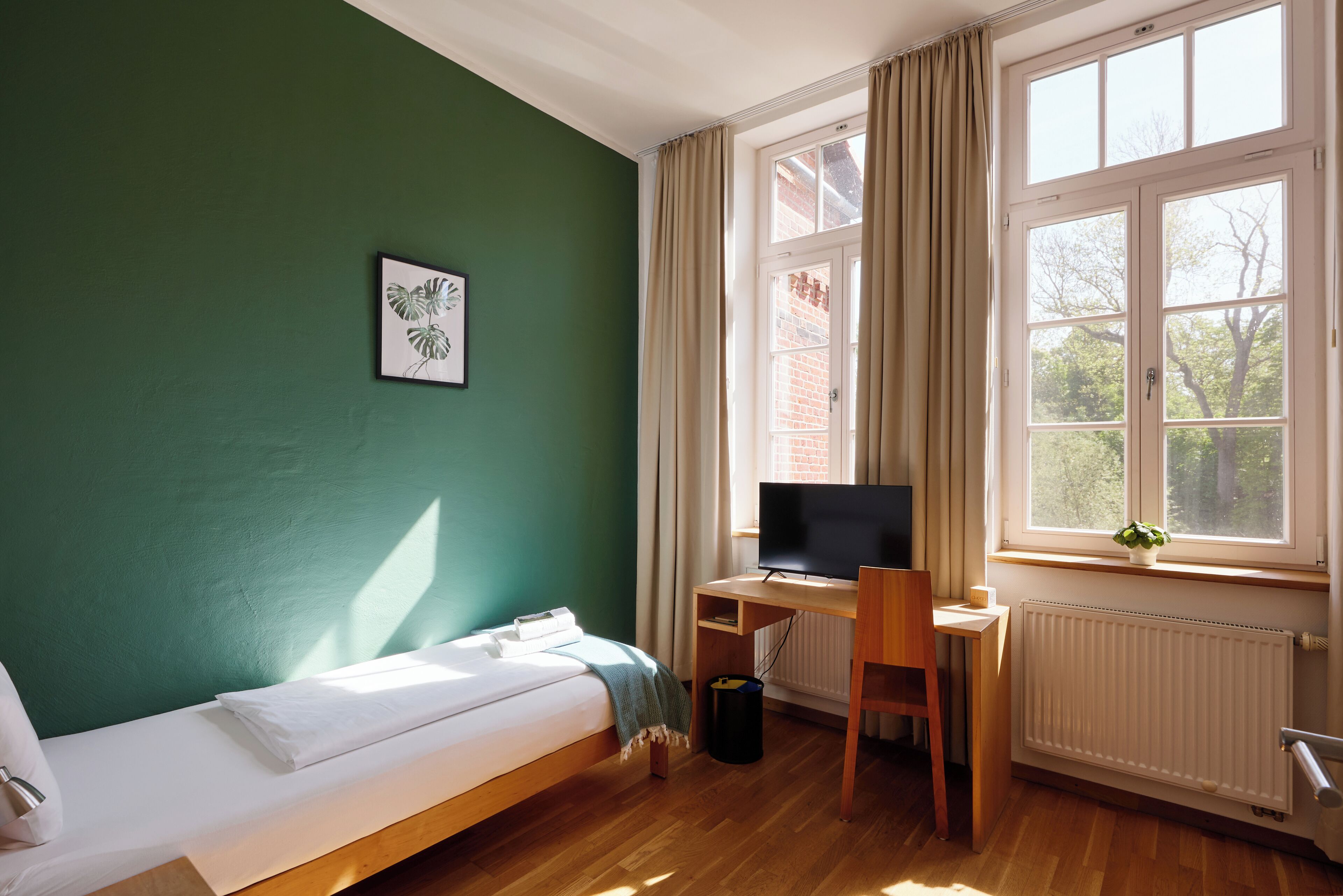 Una habitación con cama, escritorio con computadora, ventana con cortinas y radiador.