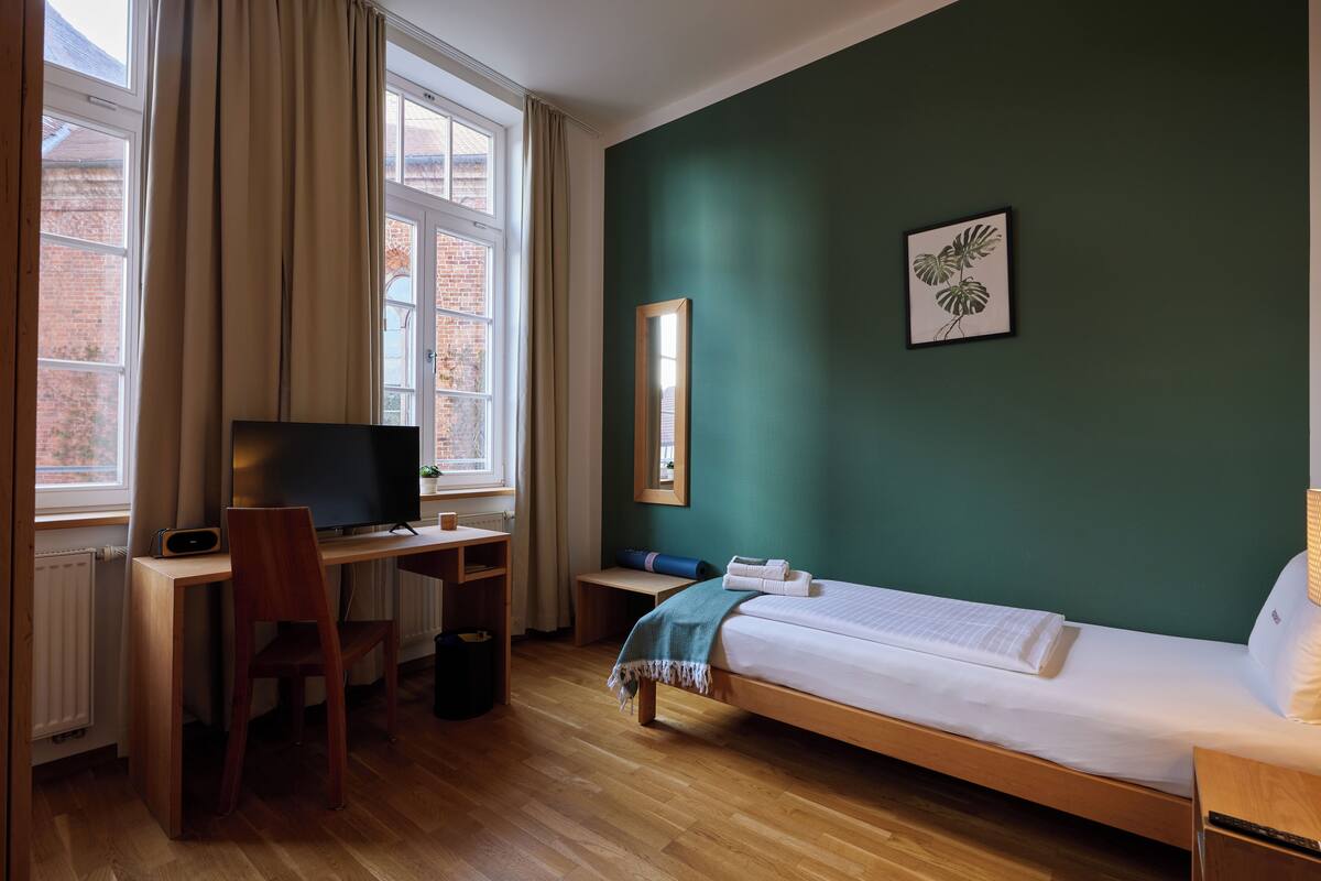 Une pièce comprenant un lit, un bureau avec un ordinateur, une chaise, un téléviseur et une fenêtre avec des rideaux.