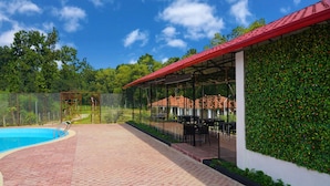 Un área de piscina con suelo de baldosas, una zona de estar cubierta con mesas y sillas, y una pared verde con plantas trepadoras.