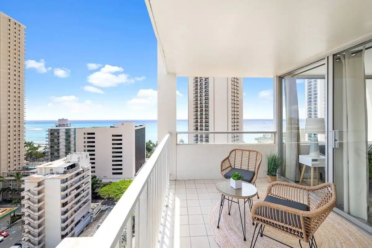 A balcony with wicker furniture, a small table, and a view of the ocean and other buildings.