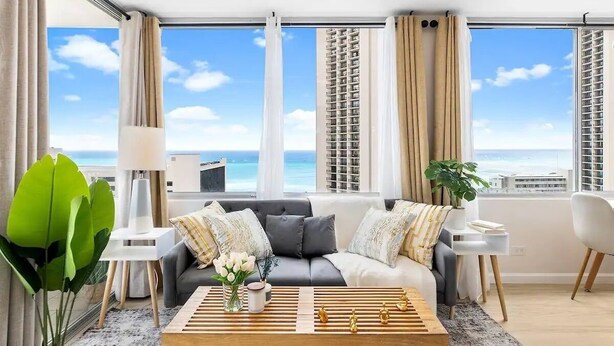 Ett modernt vardagsrum med en grå soffa, vita och mönstrade kuddar, ett träbord och utsikt över havet och höga byggnader.