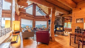 L’intérieur d’une cabane en rondins, avec une cheminée, des poutres en bois et une vue sur les montagnes.
