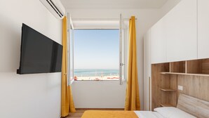 A bedroom with a bed, a TV mounted on the wall, a window with curtains, and a view of the beach. - Residence Torvaianica (Pomezia)