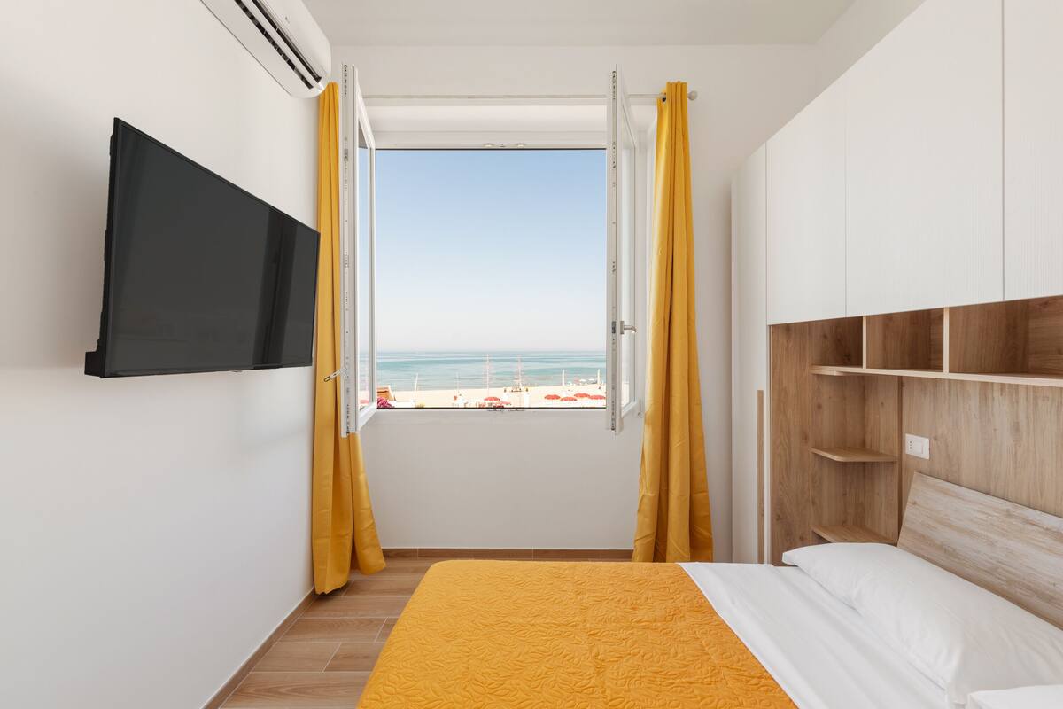 A bedroom with a bed, a TV mounted on the wall, a window with curtains, and a view of the beach.